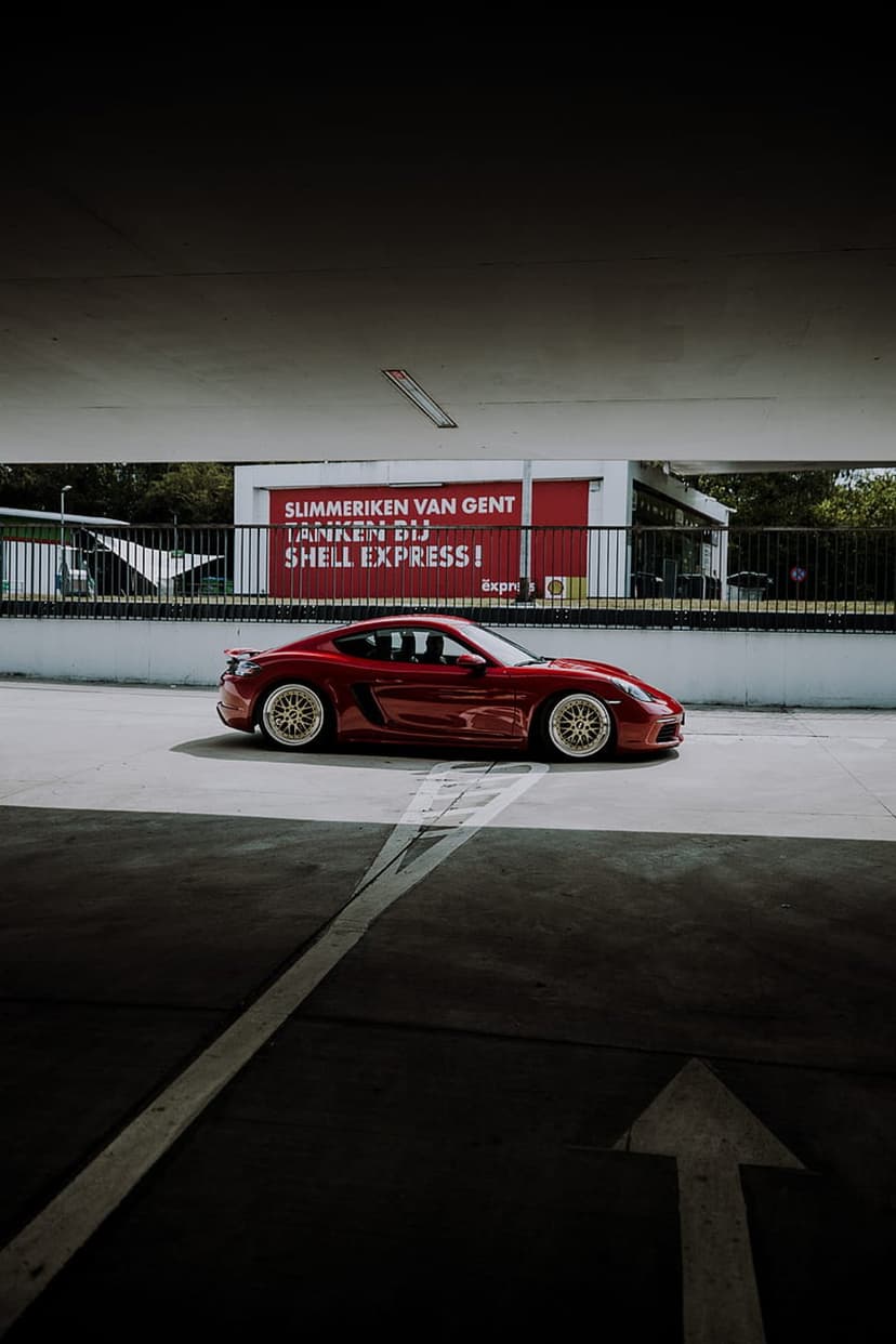 Red Porsche Cayman S with gold rims parked at a Shell Express gas station at night under a red banner.
