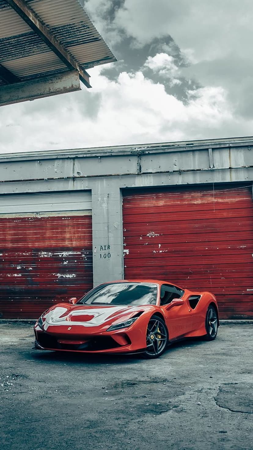 Red Ferrari F8 Tributo parked on asphalt in front of two crimson garage doors beneath a cloudy, moody sky.