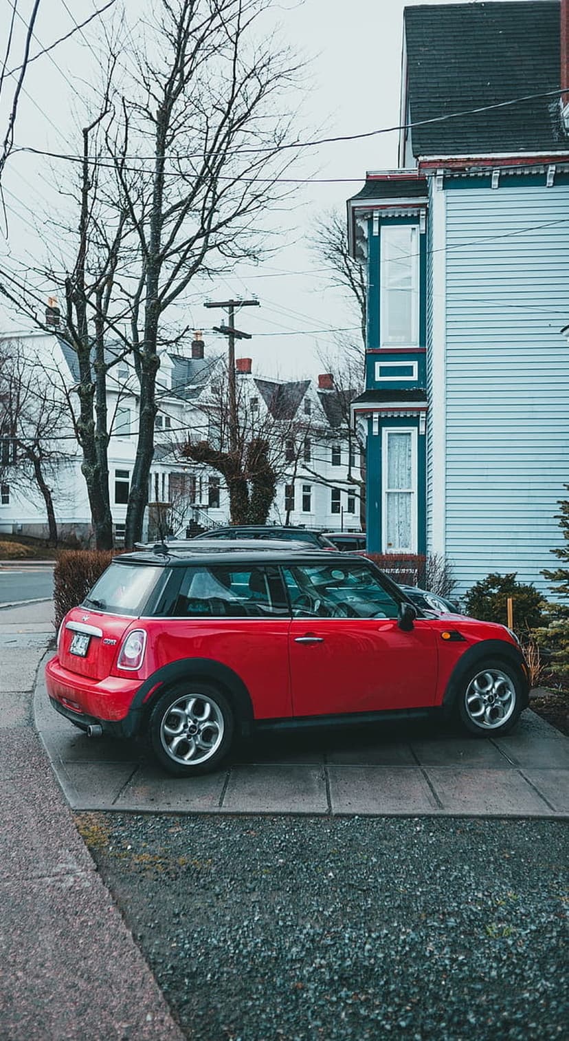 Red Mini Cooper parked on a grey tiled driveway in front of a blue suburban house with bare tree branches.