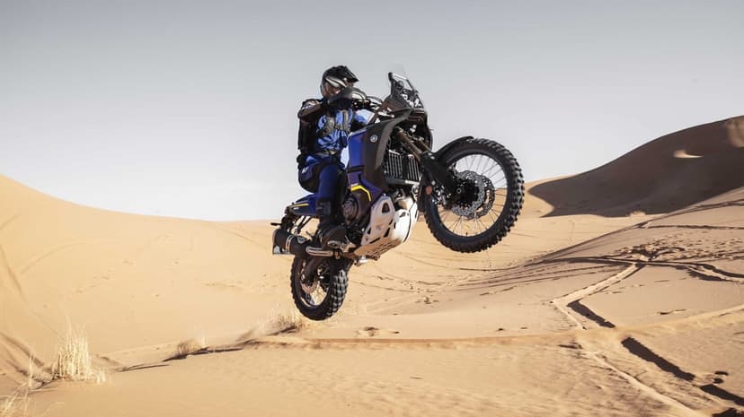Blue adventure motorcycle jumps mid-air over orange sand dunes under a bright, clear desert sky in broad daylight.