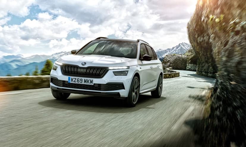 White Skoda SUV driving on a winding cliffside mountain road with snow-capped peaks under a sunny sky.