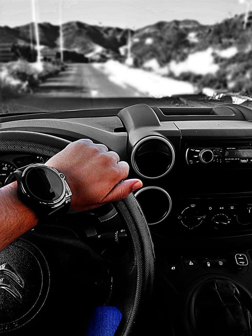 Hand wearing a smartwatch grips a car steering wheel facing a winding black and white mountain road landscape.