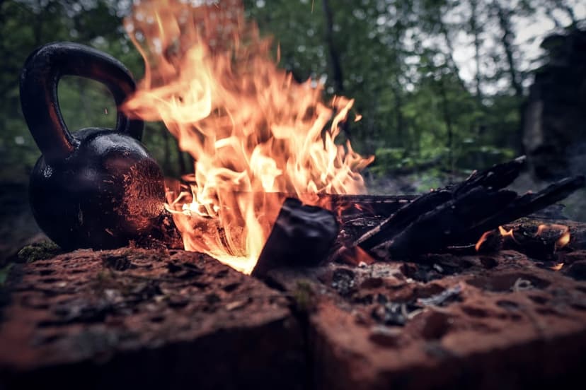 Black kettlebell rests near a brick campfire with vibrant orange flames against a blurred green forest backdrop.