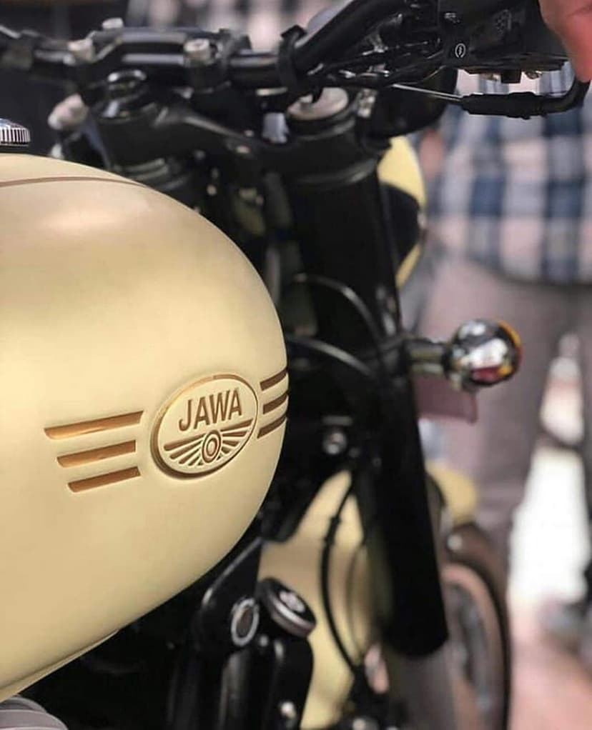 Cream Jawa motorcycle fuel tank with a raised metallic winged logo, blurred handlebars, and soft studio lighting.