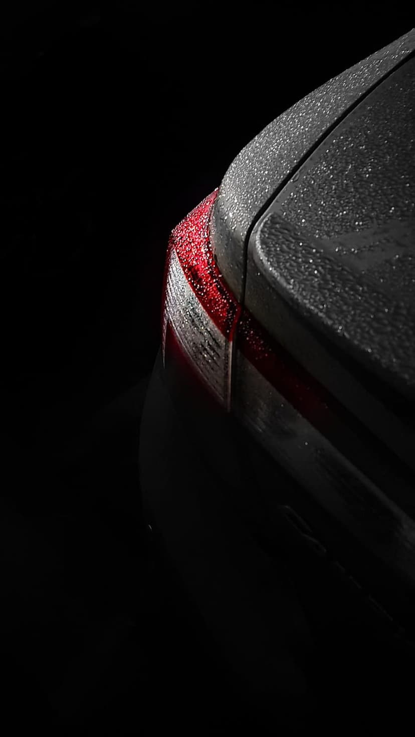 Dark car tail light covered in glistening raindrops with dramatic low-light reflections on a wet vehicle surface.
