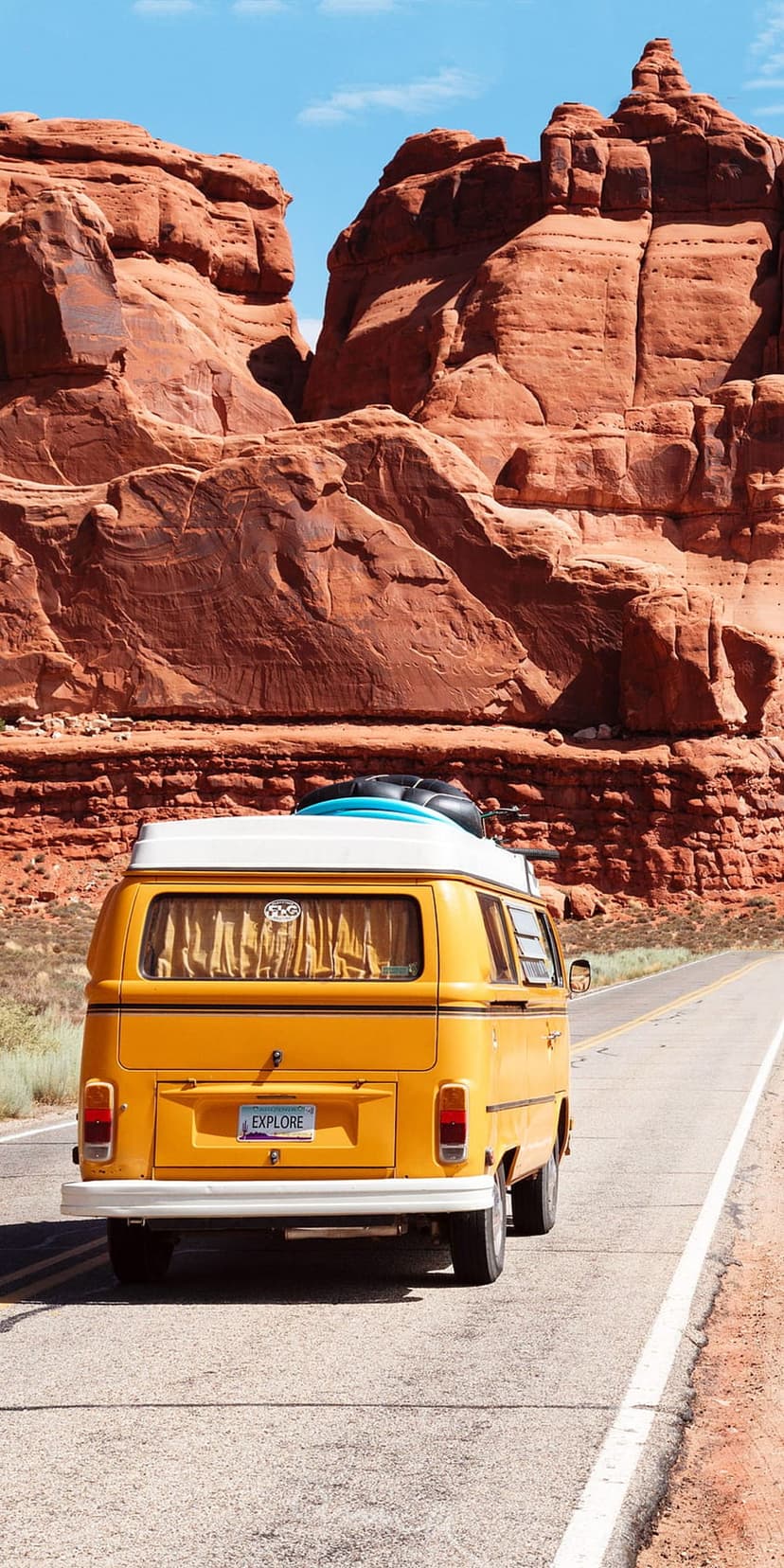 Yellow vintage van carrying surfboards travels a desert highway between towering red rock sandstone formations.