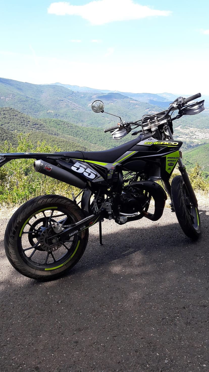 Sherco supermoto with number 55 parked on a dusty mountain road under a clear sky with rolling hills behind.