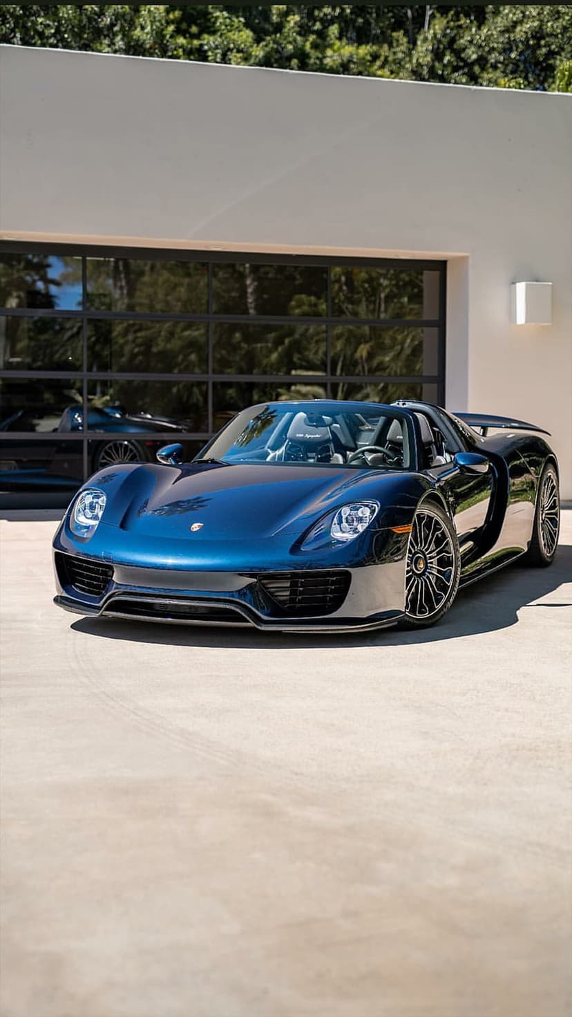 Porsche 918 Spyder in metallic blue parked before a glass garage with sky reflections on the hood and body.