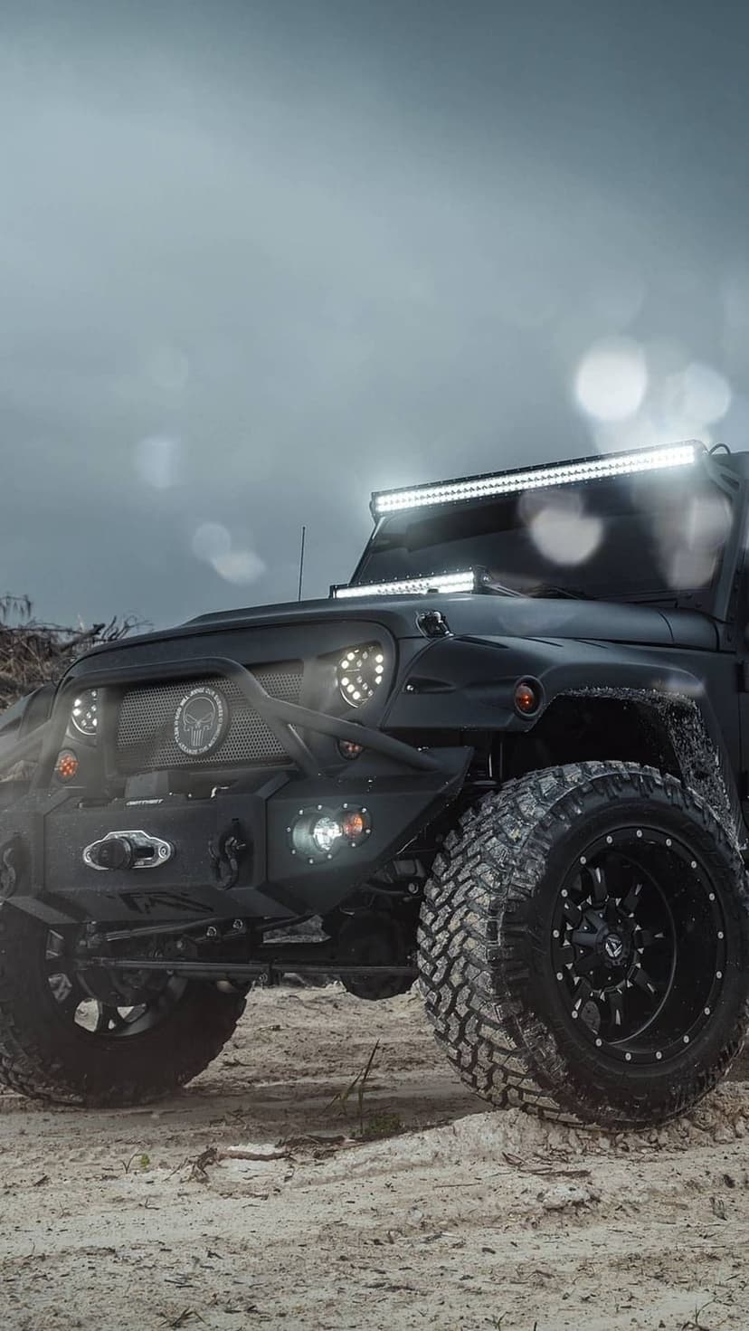 Black Jeep Wrangler with aggressive tires and LED light bars parked on sand under a cloudy, atmospheric sky.