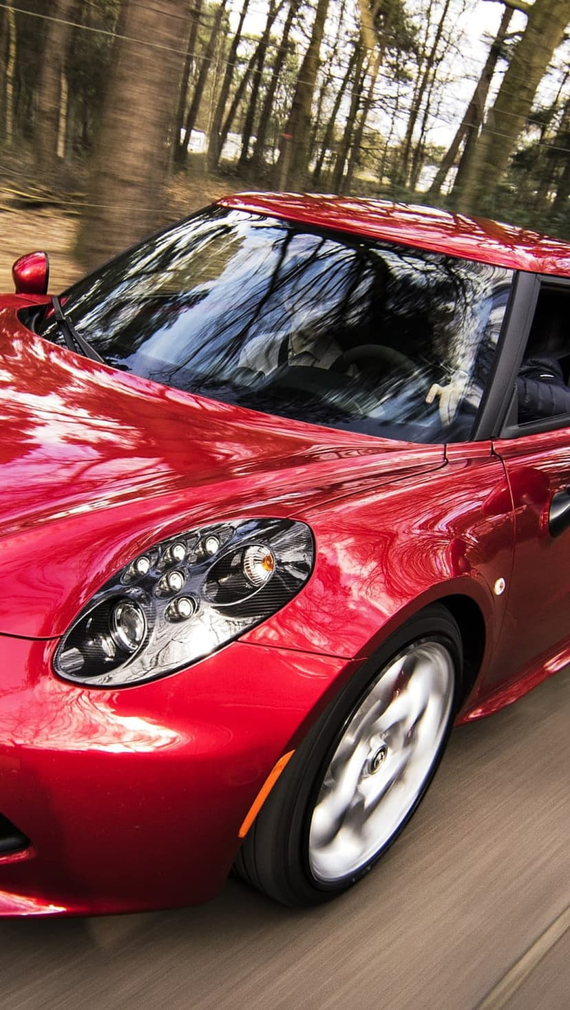 Red Alfa Romeo 4C sports car speeds along a wooded road with motion blur and glossy tree reflections.