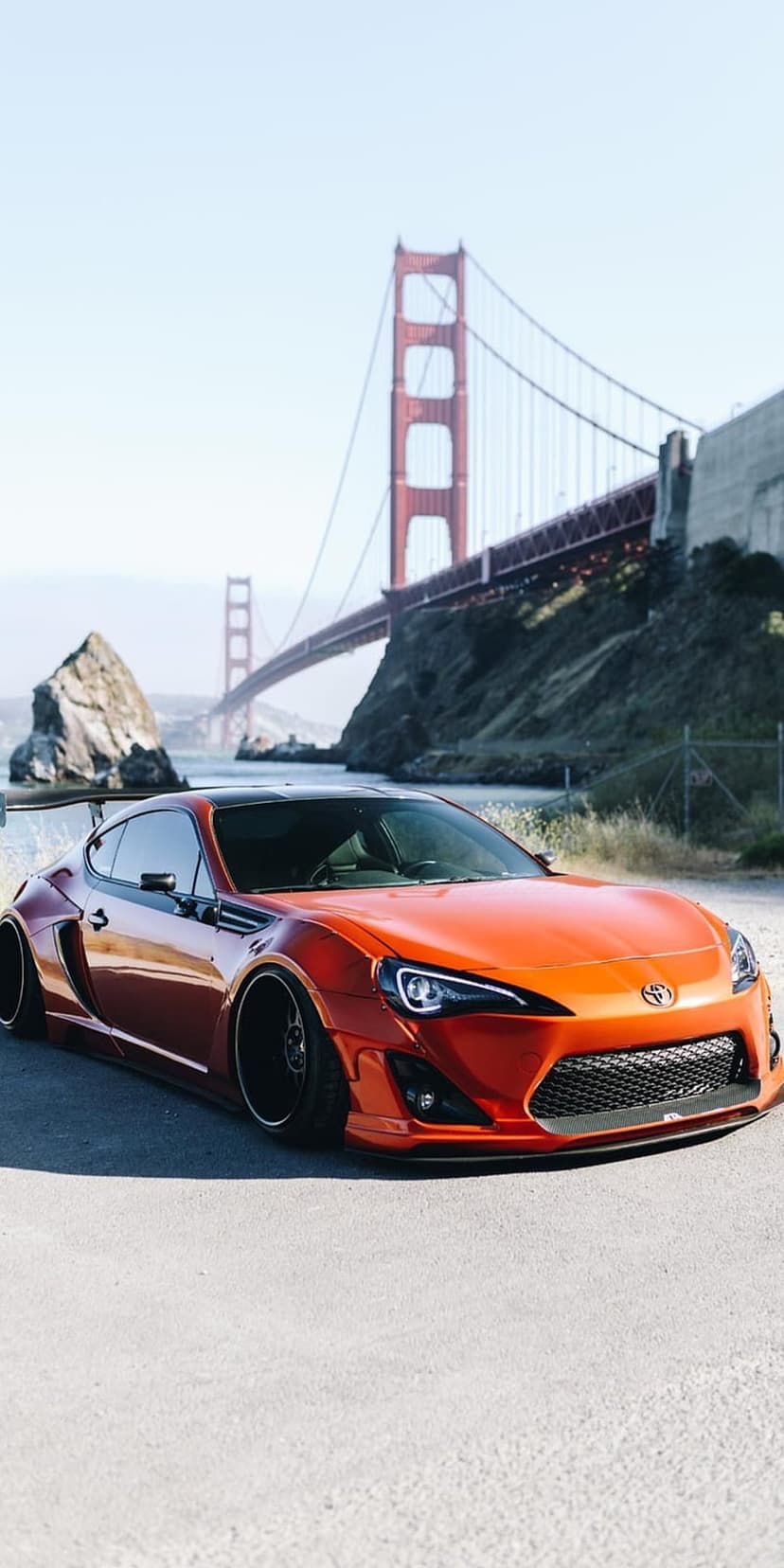Orange Toyota 86 with wide body kit parked near the Golden Gate Bridge with hills and water in the background.