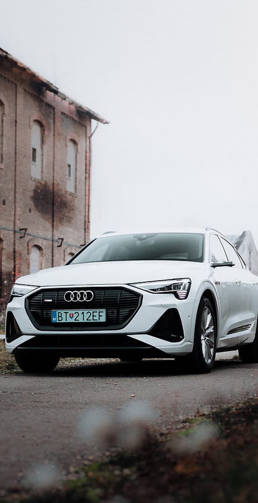 White Audi e-tron SUV parked on a paved road against an old brick building with arched windows and green plants.