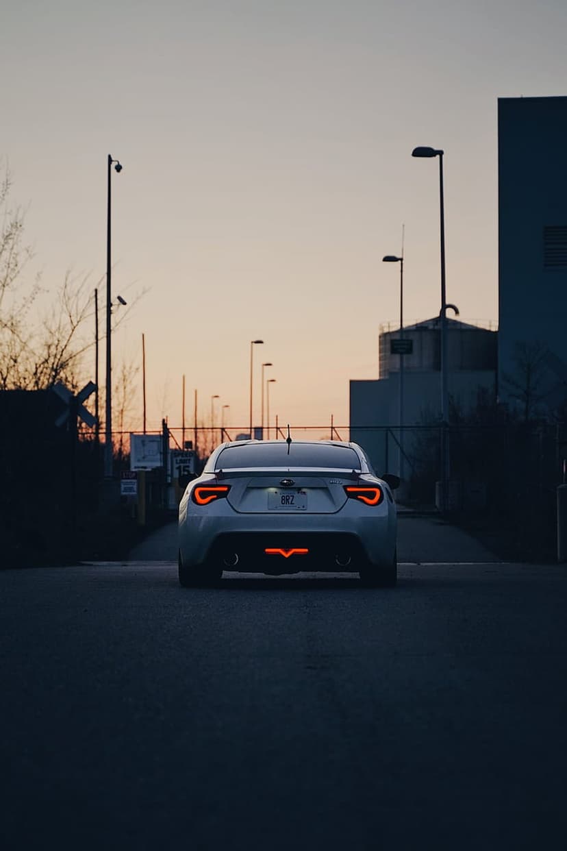 White Subaru BRZ rear view with glowing red V-shaped taillights on a dark road near an industrial building.