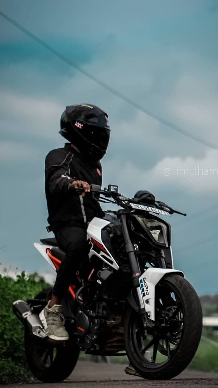 Motorcyclist in black gear sitting on a white and black KTM Duke under a cloudy blue sky with green trees.