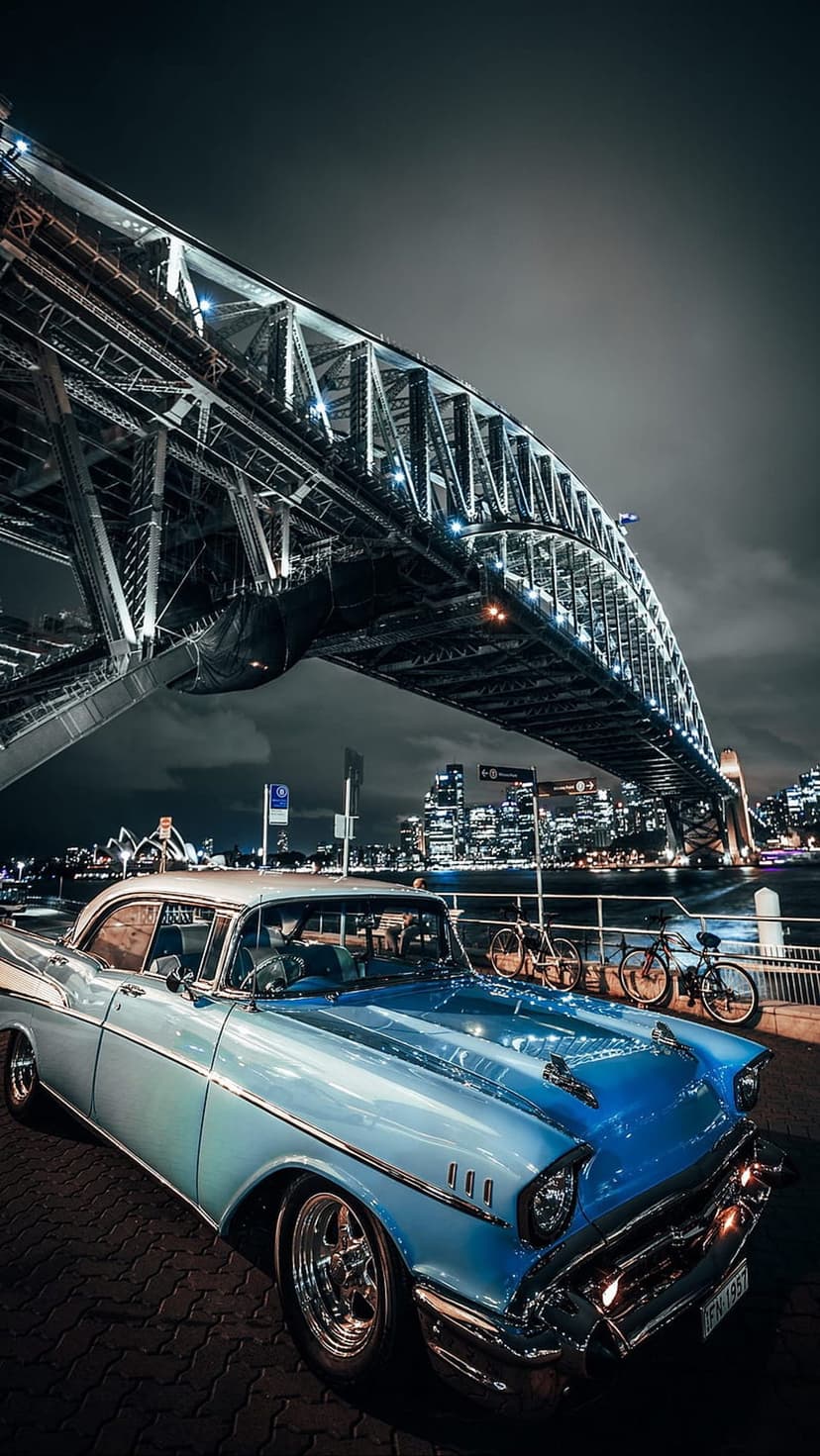 Classic blue car parked under the illuminated Sydney Harbour Bridge with the city skyline visible at night.