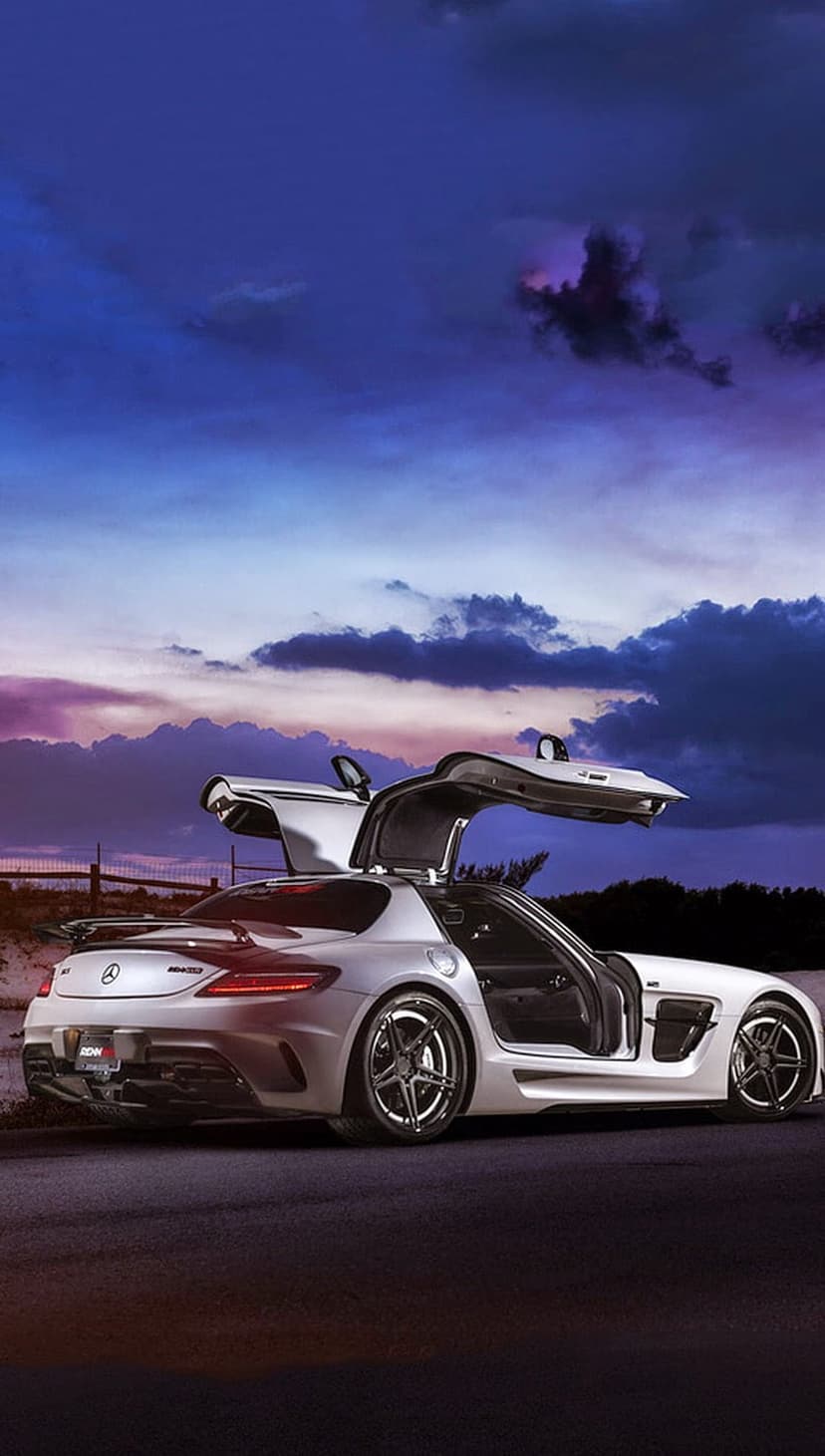 Silver Mercedes SLS AMG with open gullwing doors parked on a road beneath a vibrant purple and blue sunset sky.