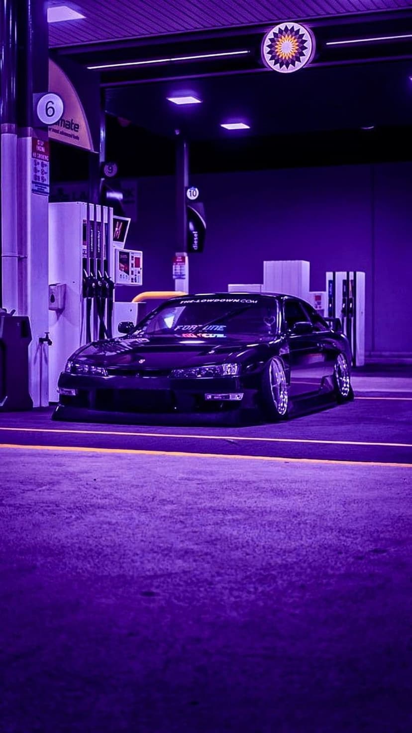Black sports car parked under purple neon lights at a gas station with reflective surfaces and fuel pumps.