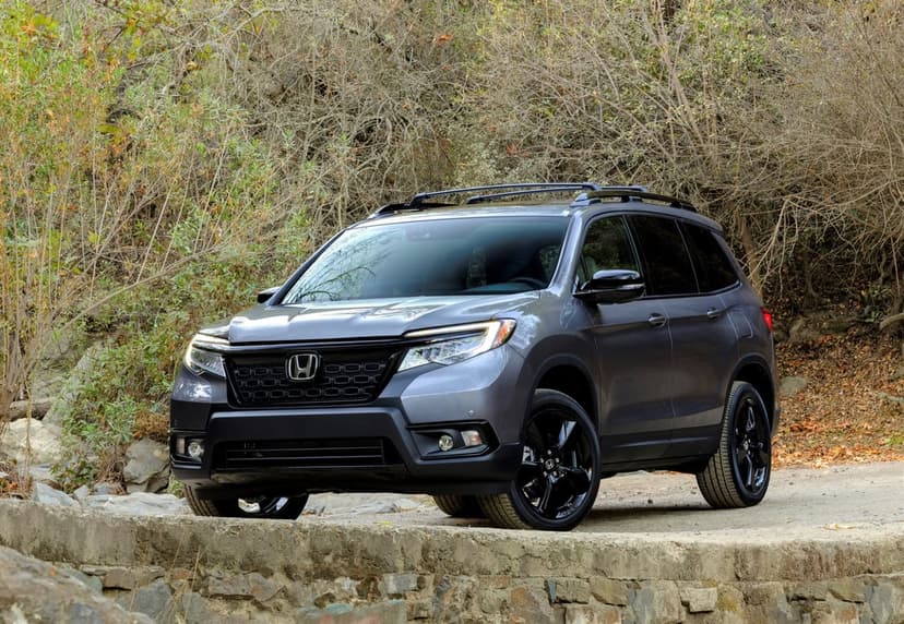 Gray Honda Passport SUV with black wheels parked on a stone bridge surrounded by a dense forest of dry trees.