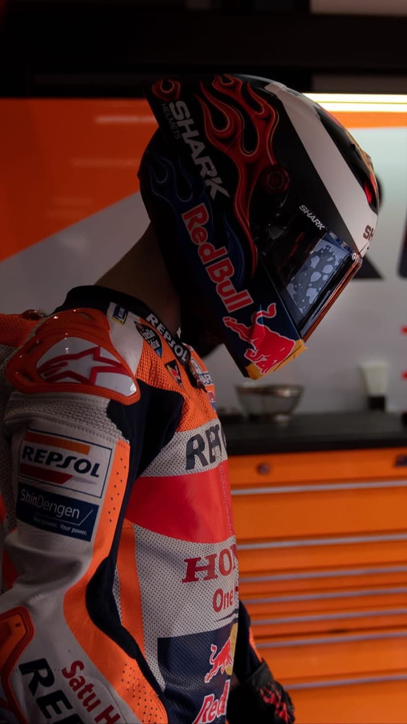 Marc Marquez wears a branded racing helmet and suit with flame graphics in a warm, orange-toned garage setting.