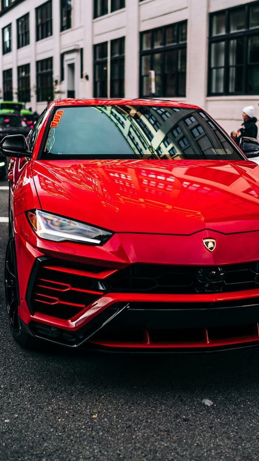 Red Lamborghini Urus SUV parked on a city street with gloss paint reflecting buildings and a person walking.