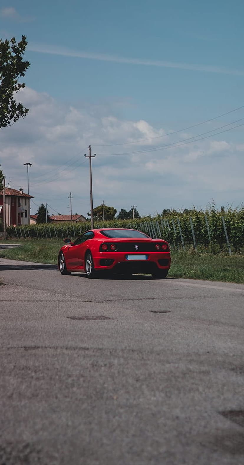 Red Ferrari sports car driving on a road past green vineyards and old Italian houses under a blue sky.