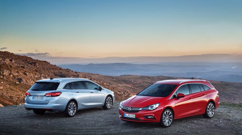 Red and silver Opel Astra Sports Tourer wagons parked on a rocky mountain ridge under a soft golden sunset sky.