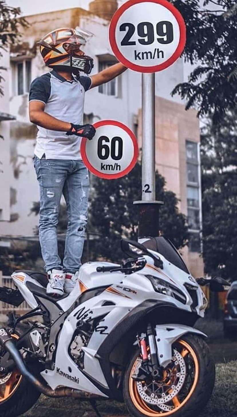 Motorcyclist in full gear stands on a white sportbike reaching for a 299 km/h speed limit sign on the road.