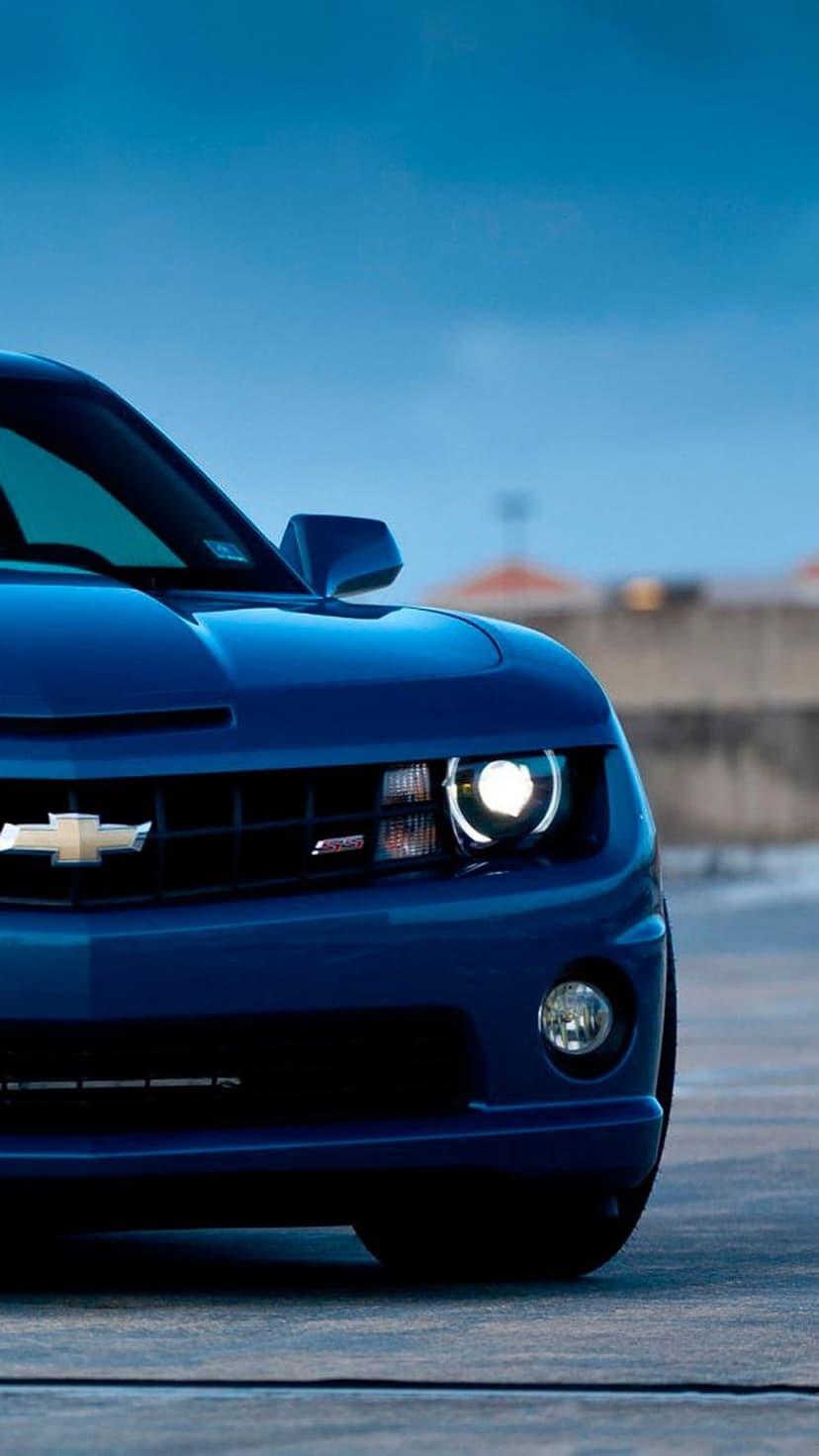 Chevrolet Camaro headlight and gold emblem on a blue car grille against a bright blue sky during daytime.