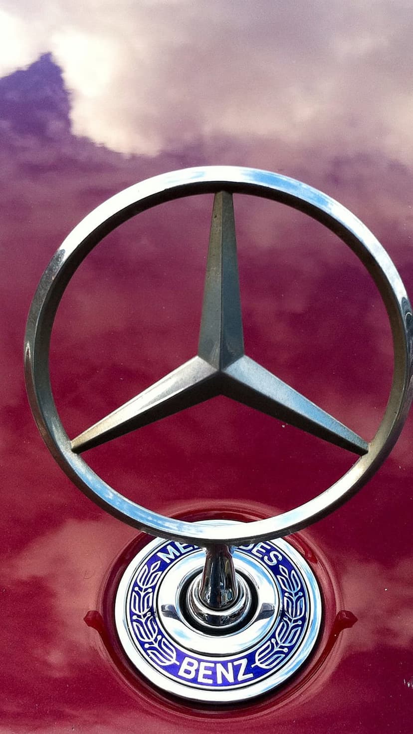 Mercedes-Benz three-pointed star logo on a red car hood reflecting a cloudy sky with purple and white hues.