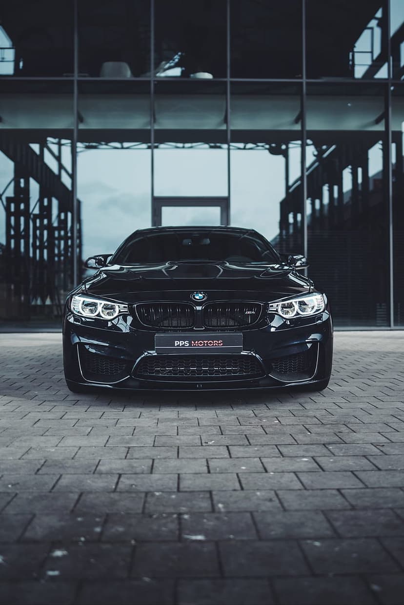 Black BMW M4 coupe parked on pavement with a modern glass building reflection and sharp LED headlights illuminated.