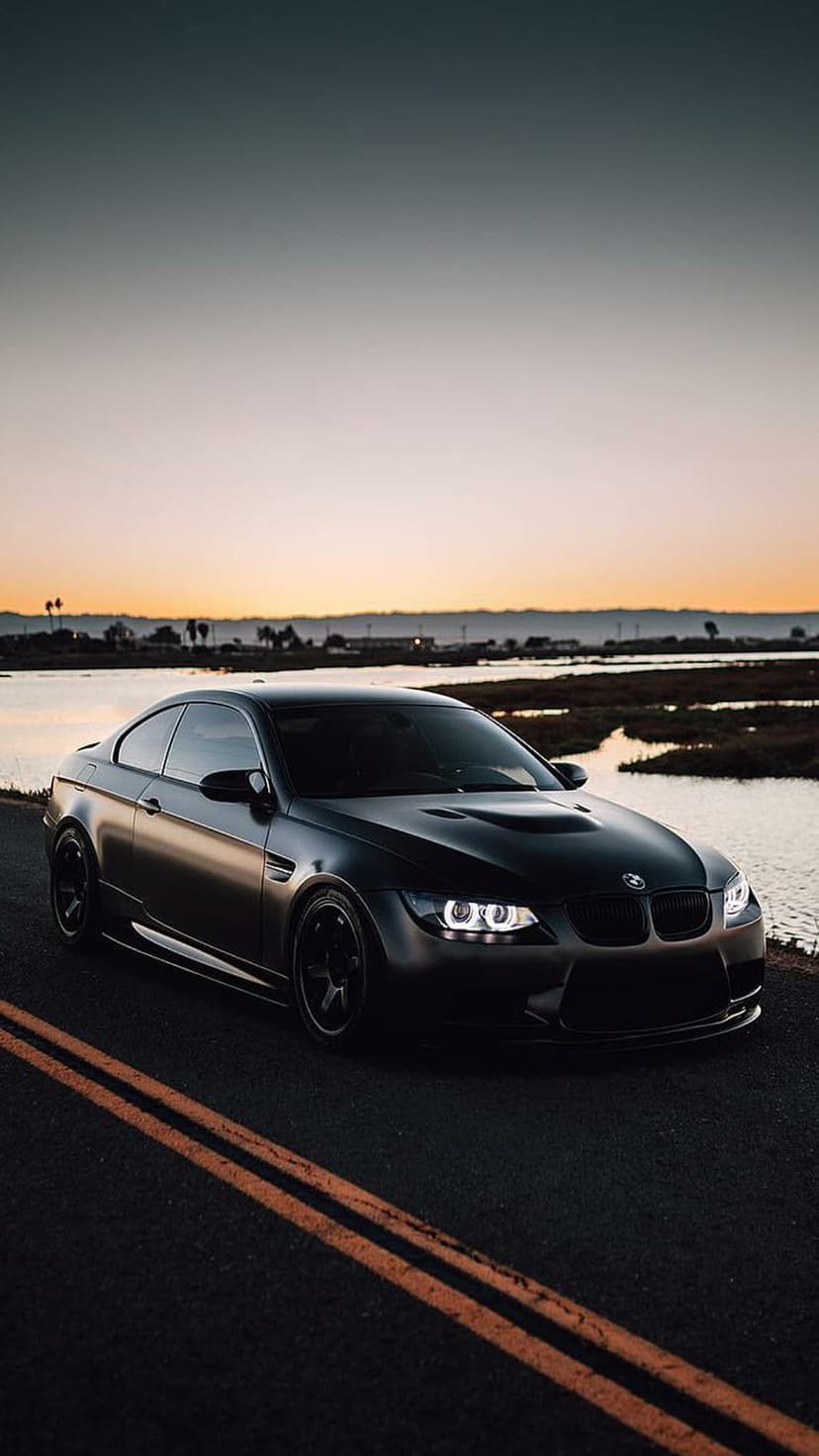 Matte black BMW M3 with glowing headlights parked on a coastal road during a vibrant orange sunset.