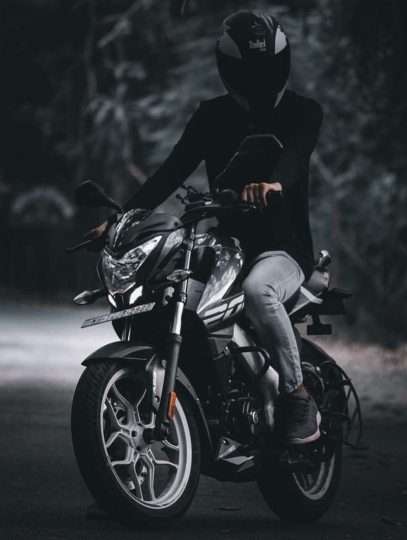 Biker in dark gear sits on a motorcycle on a paved road surrounded by a blurred, moody forest at dusk.