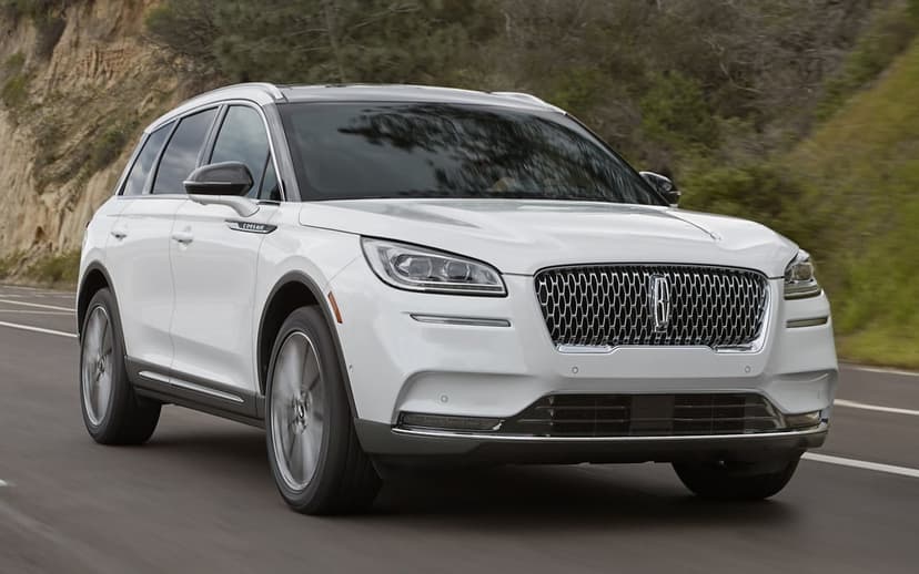 White Lincoln SUV driving on an asphalt road with a blurred green hillside background in daylight.