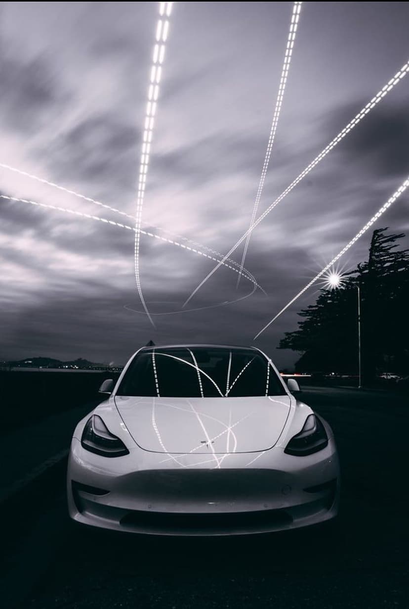 White Tesla car parked on a dark road under a cloudy sky filled with glowing neon light trails at twilight.