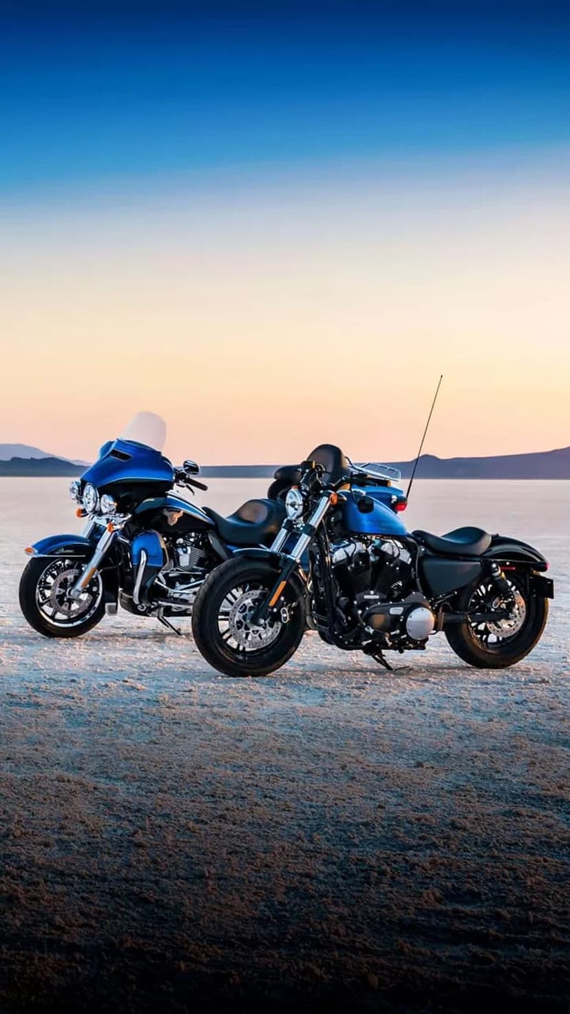 Twin blue Harley-Davidson motorcycles parked on a sandy beach under a gradient orange and blue sunset sky.