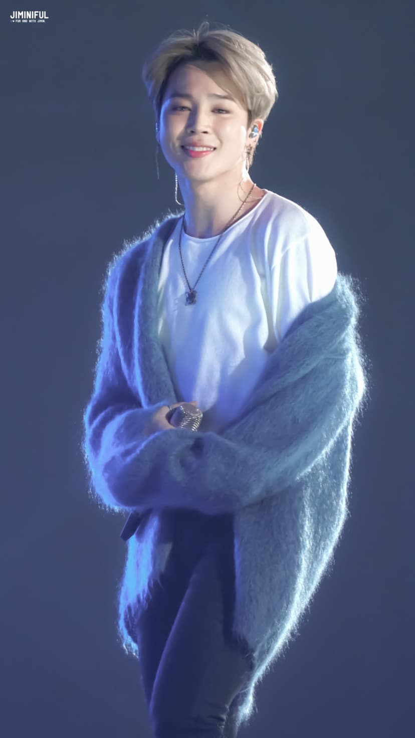 Jimin of BTS smiles with a playful expression while wearing a fuzzy blue cardigan and a thin silver necklace.