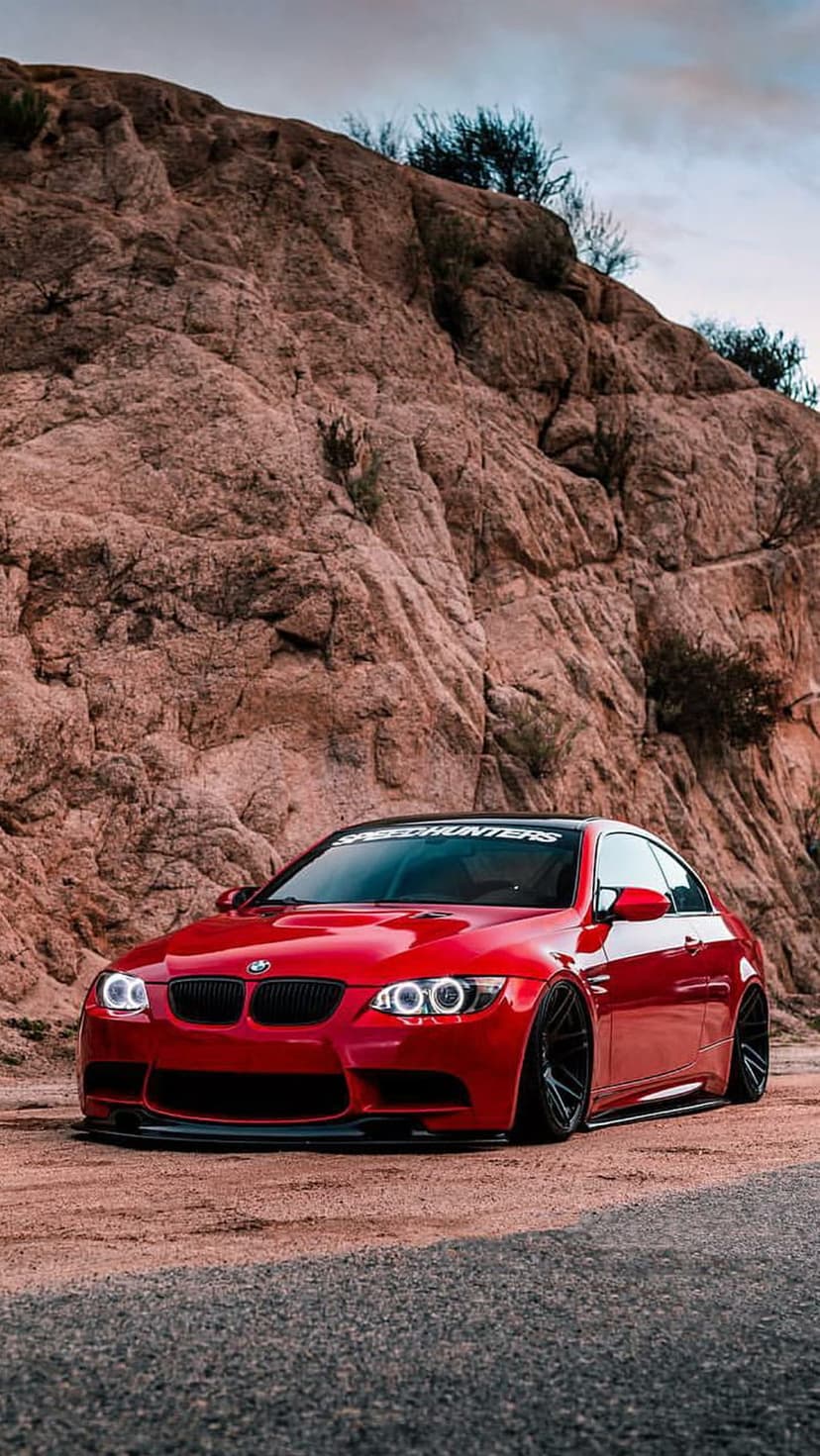 Red BMW M3 sport sedan parked on a dusty desert trail in front of a massive, textured sandstone cliff face.
