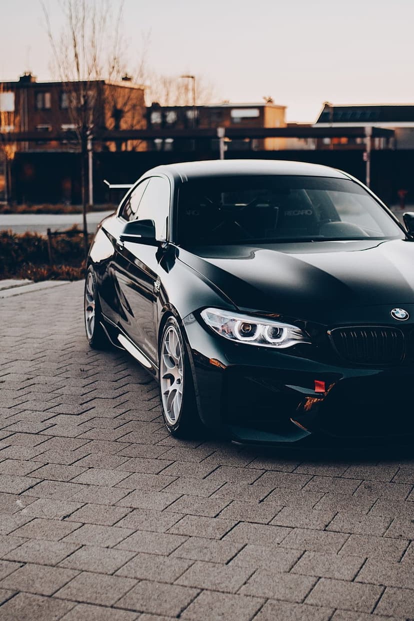 Black BMW M2 coupe on a cobblestone street at sunset with glowing headlights and a blurred urban background.