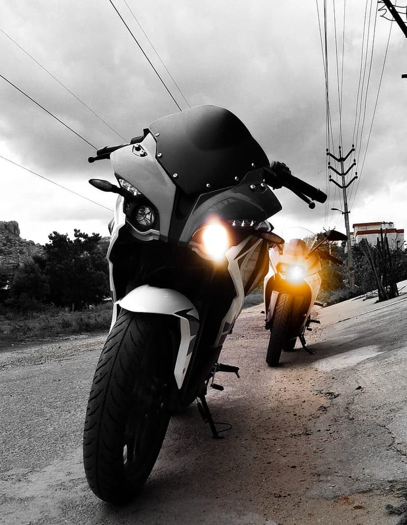 Two sport motorcycles parked on a dusty road with glowing headlights under power lines and a grey overcast sky.