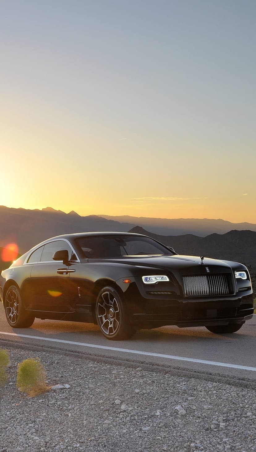 Black Rolls-Royce coupe driving on a desert road against orange sunset mountains and a golden sky background.