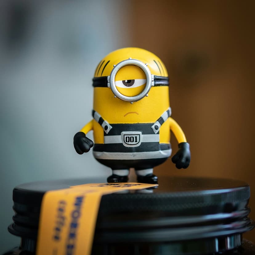 Minion toy in a striped prison uniform sits on a black jar lid with a bright yellow label in the background.