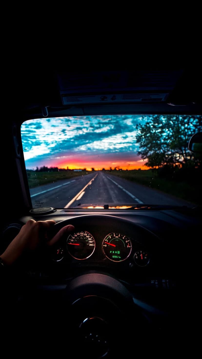Driver's hand on a steering wheel overlooking a country road and vibrant orange sunset sky with dramatic clouds.