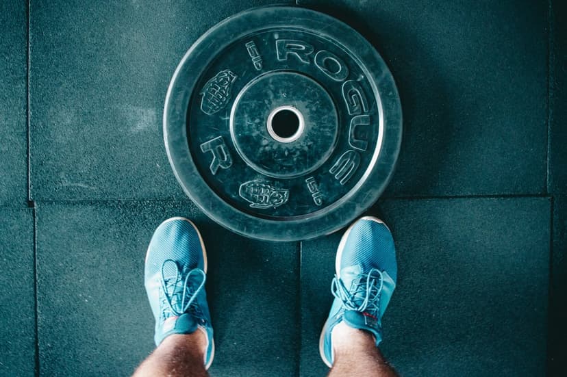 Blue athletic shoes stand on a dark gym floor beside a black Rogue weight plate during a strength workout.