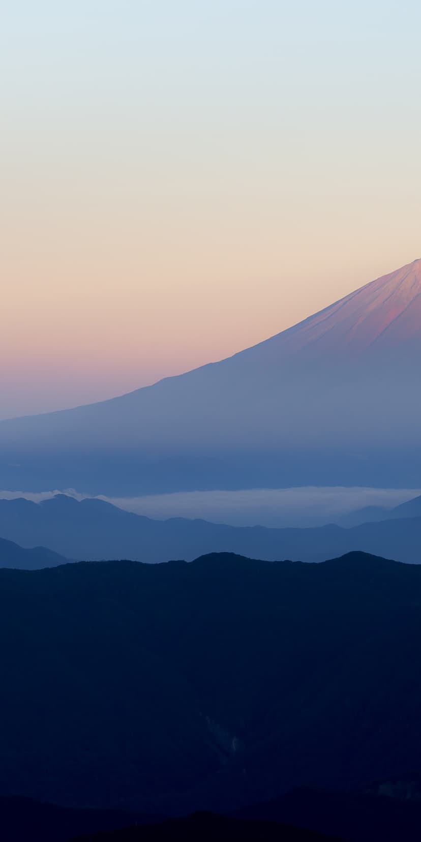 Mount Fuji summit with snow-capped peak glowing pink above layers of blue mountain ridges in the morning mist.