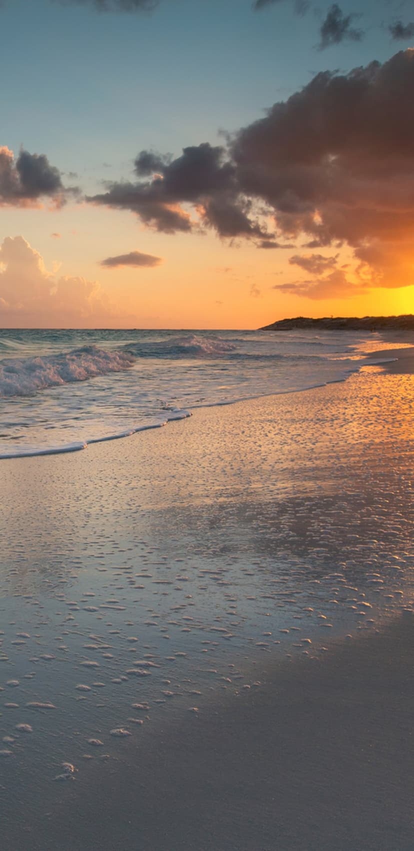Ocean waves with white foam wash over wet sand reflecting an orange and purple sunrise sky on a quiet beach.
