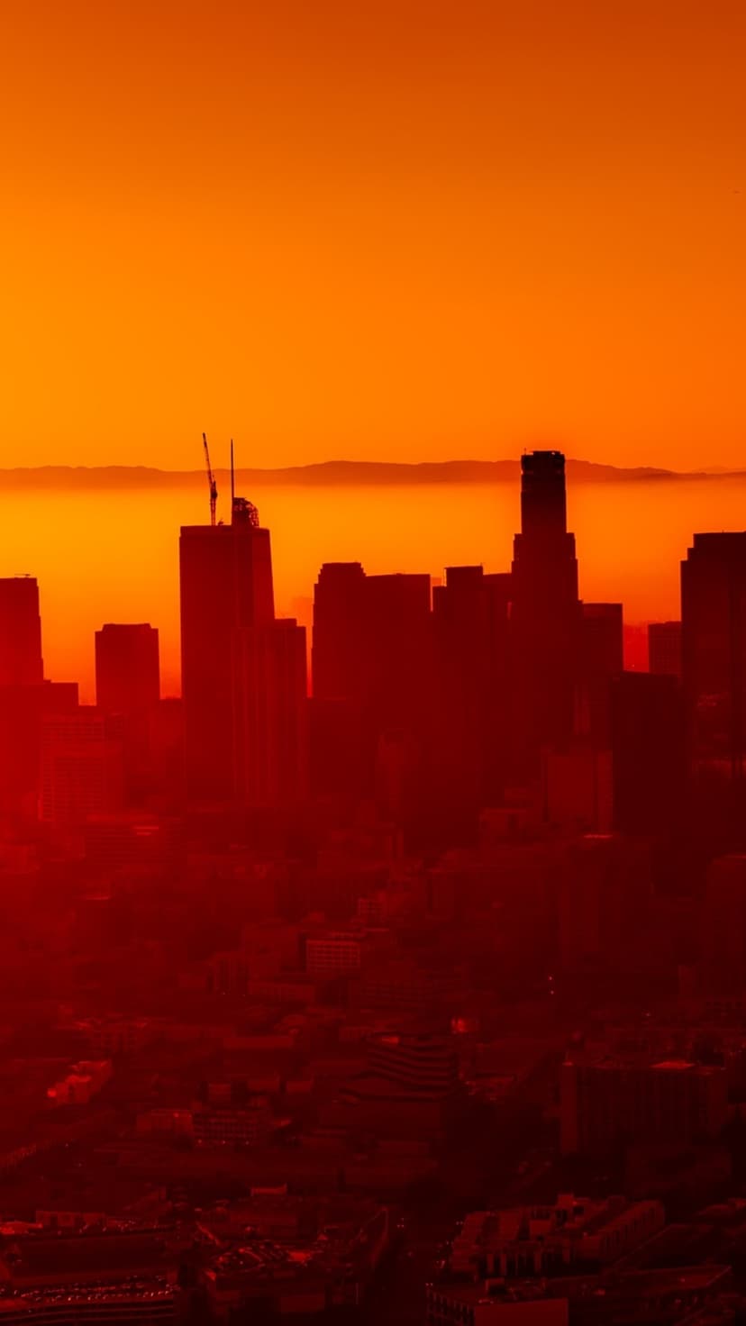 Black city buildings silhouetted against a glowing red and orange sky with hazy mountain peaks in the distance.