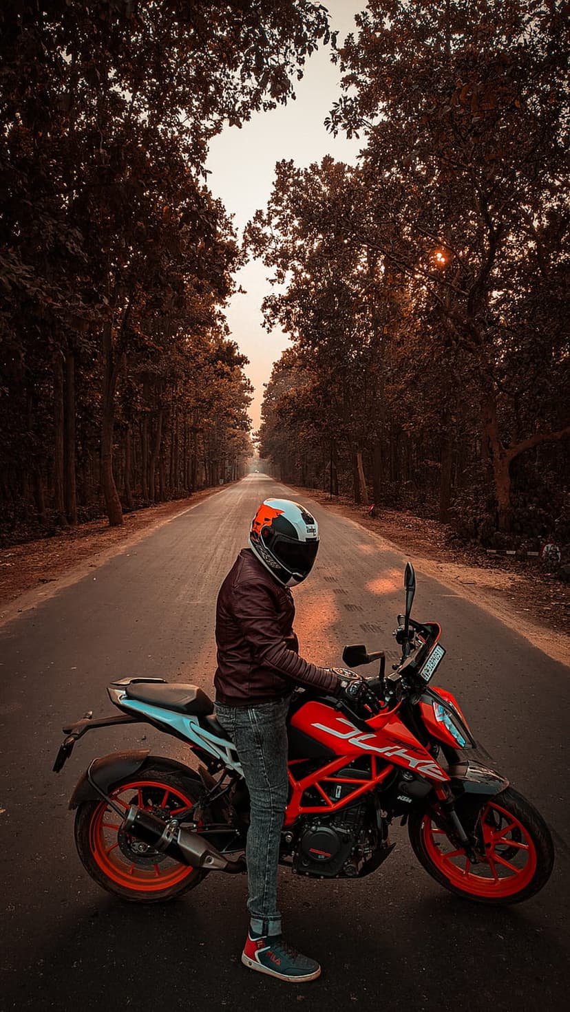 Motorcyclist in gear beside a bright bike on a forest road under a hazy orange sunset sky during autumn.