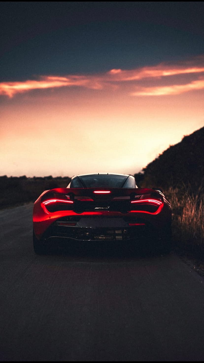 Red McLaren supercar taillights illuminate a rural asphalt road during a vivid orange and purple atmospheric sunset.