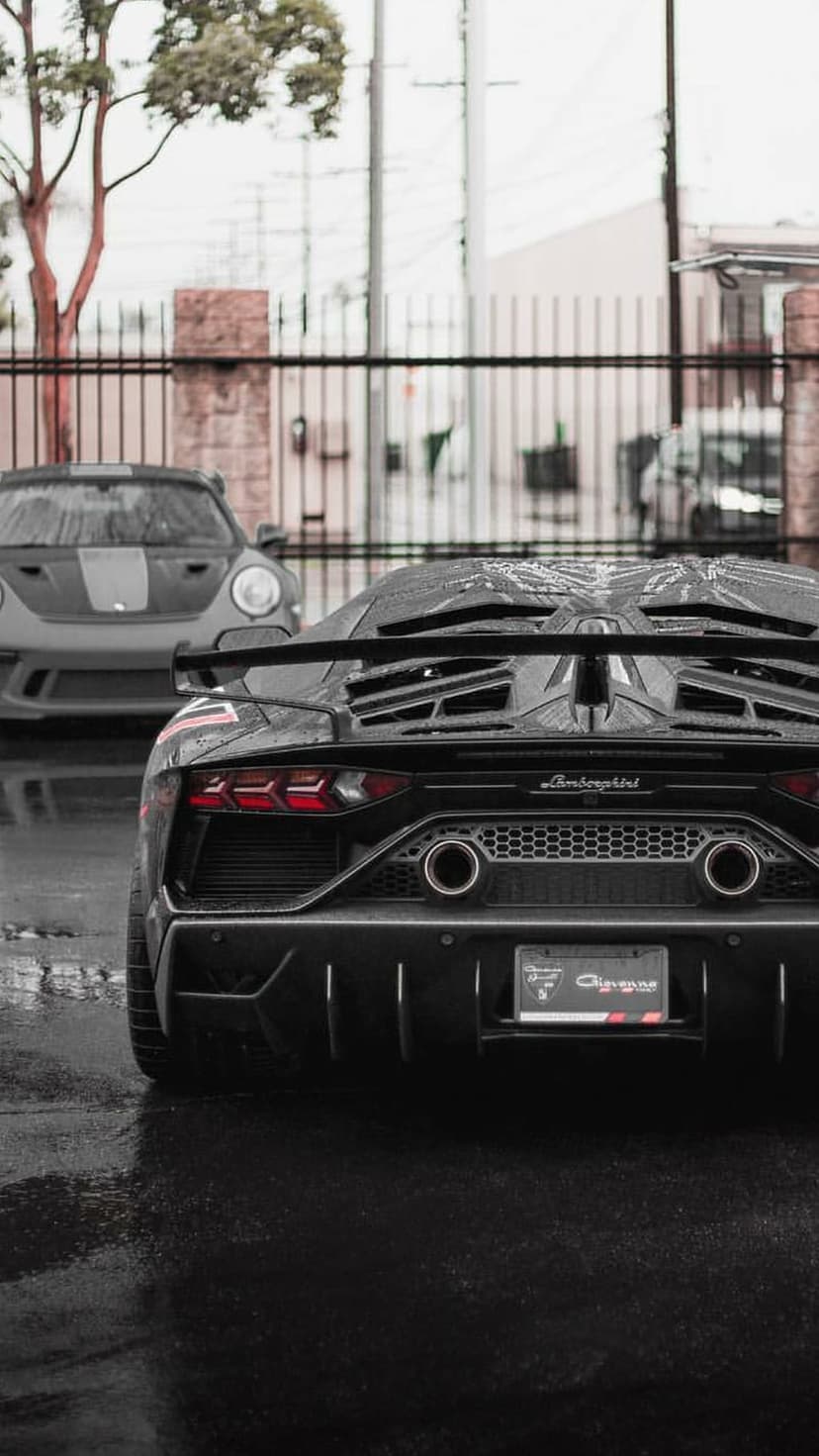 Black Lamborghini Aventador with glowing red taillights parked behind a grey Porsche 911 on a wet asphalt street.