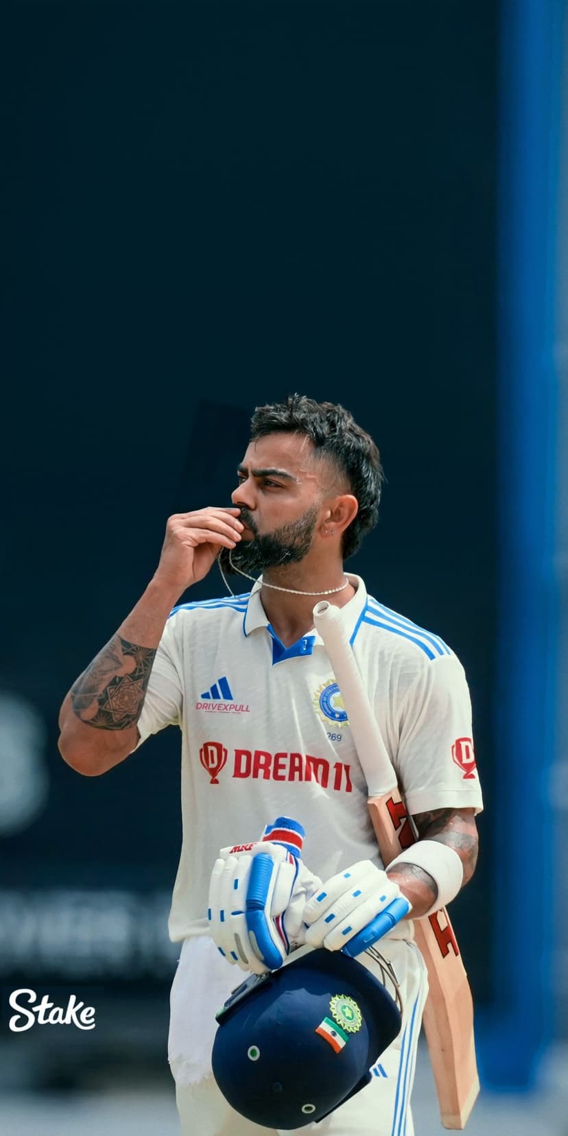 Virat Kohli wears a white Test cricket jersey and pearl necklace while holding a cricket helmet and bat.